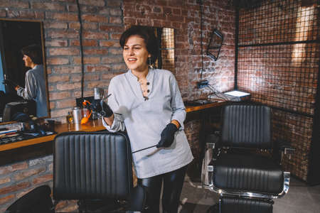 Young beautiful hairdresser woman with scissors in hand against a brick wall background. Barbershop advertising concept. Place for text or advertising.の写真素材