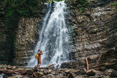 Traveler man with a yellow backpack standing on the background of a waterfall makes a photo landscape. Travel lifestyle concept. Copy, empty space for text.の写真素材