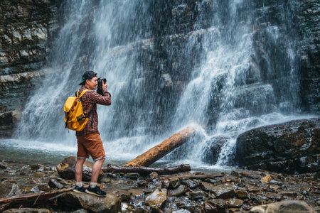 Traveler man with a yellow backpack standing on the background of a waterfall makes a photo landscape. Travel lifestyle concept. Copy, empty space for text.の写真素材
