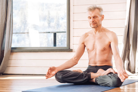 Caucasian senior man practicing yoga classic asana in studio, training in front of a window. Copy, empty space for text.の写真素材