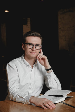 Young man wearing white shirt and eye glasses sitting at the table holding pen takes notes to his...