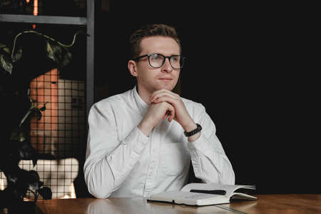 Serious young man wearing white shirt and eye glasses sitting at the table holding pen takes notes to his agenda on a dark background in office. Copy, empty space for text.の写真素材