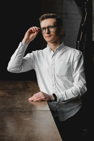 Serious young man wearing white shirt and eye glasses sitting at the table holding pen takes notes to his agenda on a dark background in office. Copy, empty space for text.の写真素材