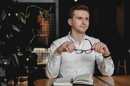 Serious young man wearing white shirt and eye glasses sitting at the table holding pen takes notes to his agenda on a dark background in office. Copy, empty space for text.の写真素材