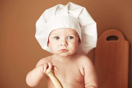 Little boy baker in a chef's hat sitting on the table playing with flour on a brown background with a wooden rolling pin, a round rustic sieve and eggs. Copy, empty space for text.の写真素材