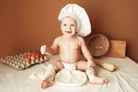 Little boy baker in a chef's hat sitting on the table playing with flour on a brown background with a wooden rolling pin, a round rustic sieve and eggs. Copy, empty space for text.の写真素材