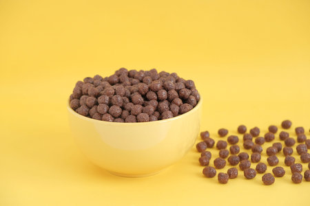 Chocolate cereal corn balls in a yellow bowl scattered on a yellow background. Copy, empty space for...