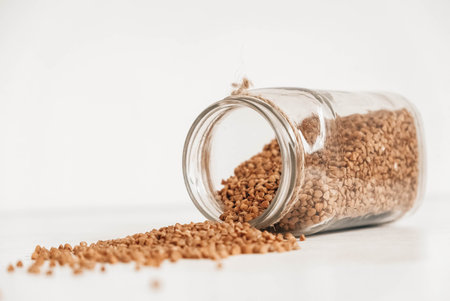 Buckwheat in a glass jar scattered on a wooden white table. Copy, empty space for text.