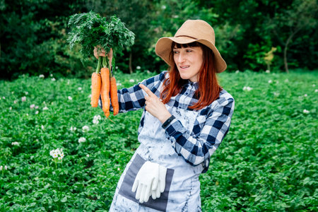 Portrait of a beautiful woman farmer holds a bunch of carrots in a straw hat and surrounded by the many plants in her vegetable garden.の写真素材