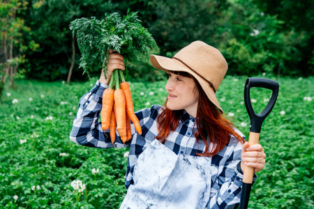 Portrait of a beautiful woman farmer holds a bunch of carrots in a straw hat and surrounded by the many plants in her vegetable garden.の写真素材