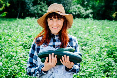 Portrait of a beautiful woman farmer holds a bunch of carrots in a straw hat and surrounded by the many plants in her vegetable garden.の写真素材