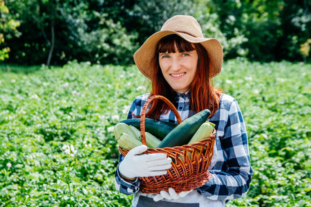 Portrait beautiful female farmer holds a zucchini in a wicker basket wearing a straw hat and surrounded by the many plants in her vegetable garden.の写真素材