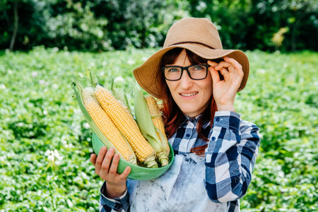 Portrait of a beautiful female farmer holding a corns cobs in a plate, smiling at the camera in a straw hat and surrounded by plenty of plants in her vegetable garden.の写真素材