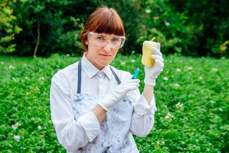 Scientist biologist in a lab coat and protective gloves introduces a blue liquid into the vegetables, yellow fresh bell pepper, against the background of green plants. Genetic breeding of vegetables.の写真素材