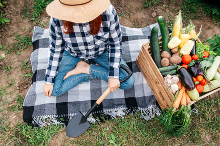 Beautiful female farmer sitting on a blanket near fresh organic vegetables in a wooden box against the background of a vegetable garden.の写真素材