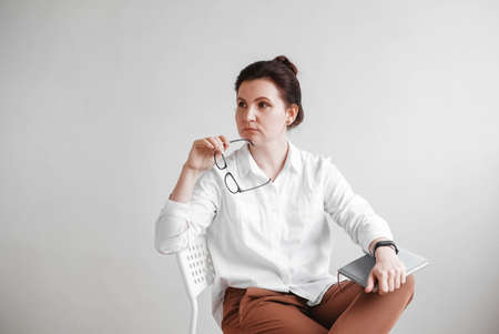 Woman in optical glasses and a white shirt sitting on a chair on a white background. Copy, empty space for text.の写真素材