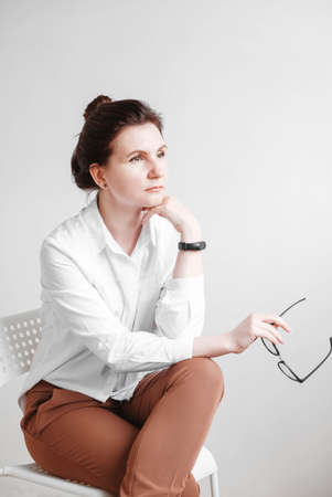 Woman in optical glasses and a white shirt sitting on a chair on a white background. Copy, empty space for text.の写真素材