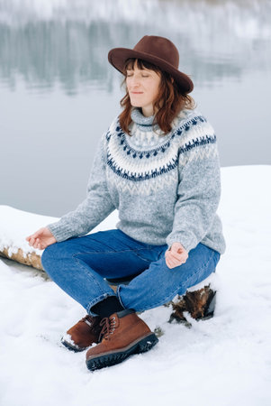 Woman is sitting and meditating in the yoga pose on the snow on a background of winter landscape.