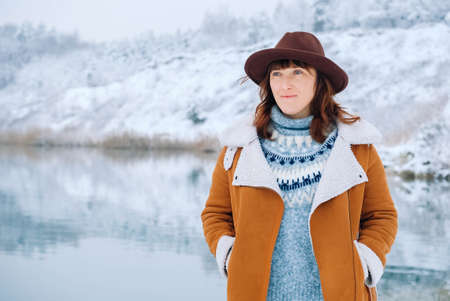 Woman wearing hat and warm jacket standing on the shores of a snow-covered lake and forest.の写真素材