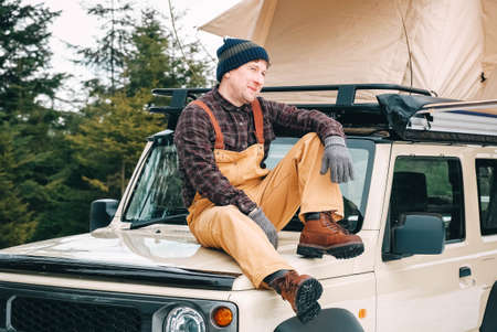 Adventurer man sitting down on the roof of the car with tent mounted on it for free travel camping lifestyle.の写真素材