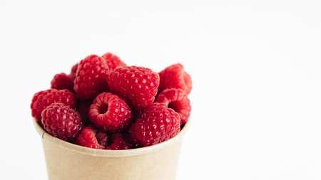 Fresh red raspberries in a paper cup on a white table background.