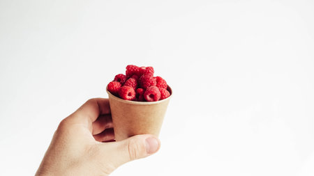 Fresh raspberries in a paper cup held by a male hand on a white background.