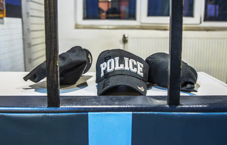 Three black police caps including one featuring bold white embroidered text are displayed on a white surface behind dark vertical bars suggestive of confinement or display.の写真素材