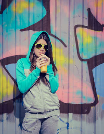 Stylish teenage girl in colorful sunglasses drinking juce near graffiti wallの写真素材