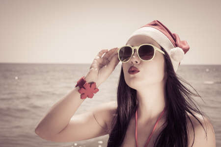 Funny young woman  in Santa Claus hat and sunglasses on a beach. Retro style toned effectの写真素材