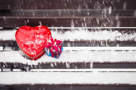 Red heart shaped tin box and a gift box on a bench covered with snow. Valentine's day themeの写真素材