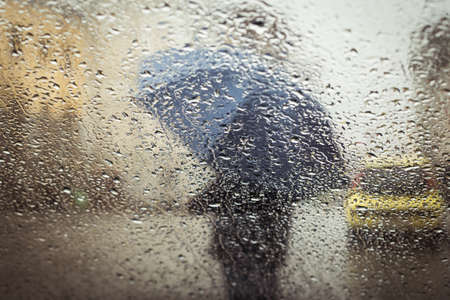 A blurry silhouette with umbrella on a city street seen through wet windowの写真素材