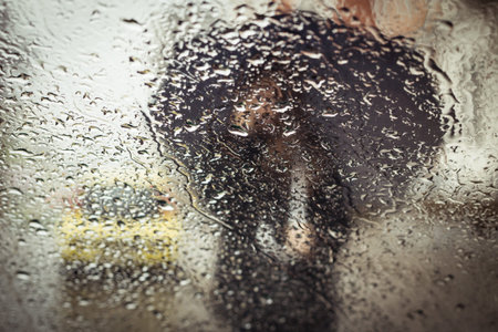 A blurry silhouette with umbrella on a city street seen through wet window