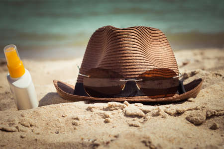 A hat with sunglasses and a bottle of sunscreen lotion lying on the sand. Vacation theme. Toned effectの写真素材