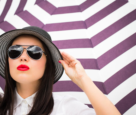 Fashion portrait of a young woman in a hat and sunglasses next to striped background.