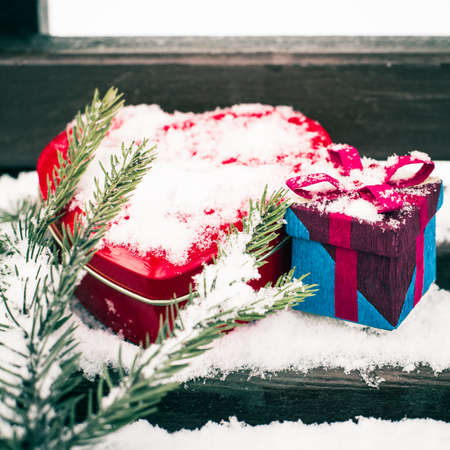 A gift box with red heart shaped tin box and a fragment of a pine-tree branch on a bench covered with snowの写真素材