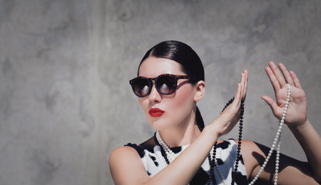 Stylish beautiful woman with sunglasses, pearl necklaces and  bright painted lips posing next to...