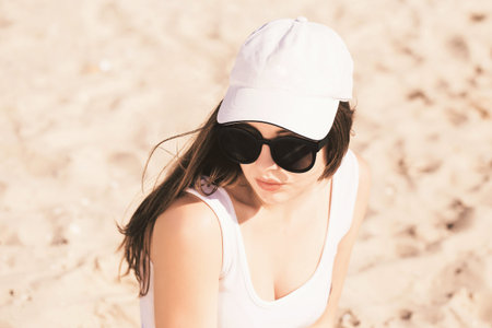 Portrait of a pretty teenage girl with long hair wearing white baseball cap, white swimsuit and...