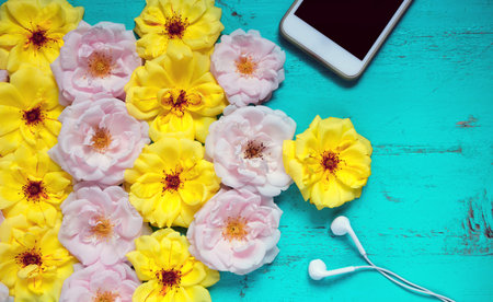 Beautiful summer background with fresh roses, smartphone and white earphones on an old painted...