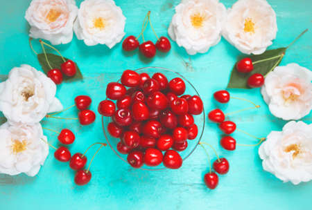 Fresh red cherry in a bowl on an old painted wooden table with fresh white roses as a bright colorful summer background for seasonal cards, posters, blogs, web designの写真素材