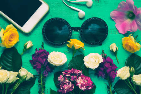 Trendy sunglasses, smartphone, earphones and summer flowers on an old painted wooden table.Bright set of fashion accessories. Colorful summer background for blogs, postrers, cards and web design. Top view. Flat layの写真素材