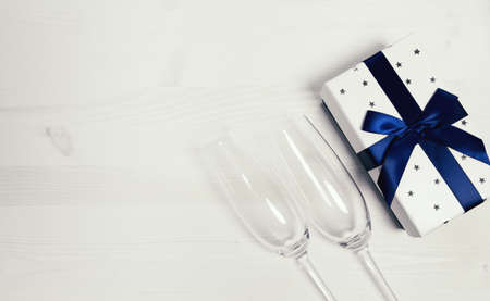 Two chrystal champagne glasses and a gift box tied with a blue ribbon on a white painted wooden table.  Celebration background. Holiday and festive backdrop. Flat lay style. Christmas or birthday gift. Top view. Copy space.の写真素材