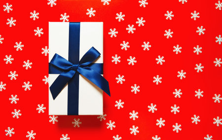White present box with dark blue ribbon and bow on red background with decorative snowflakes....
