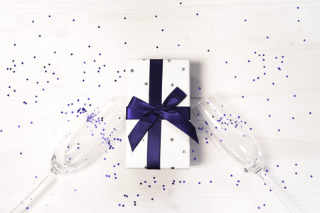 Two chrystal champagne glasses and a gift box tied with a blue ribbon on a white painted wooden table. Holiday and festive backdrop. Flat lay style. Christmas or birthday gift. Top view. Copy space.の写真素材