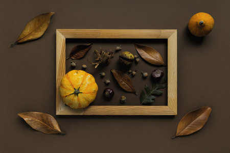 Autumn composition made of pumpkins, dried leaves, chestnuts and acorns in a wooden frame on brown...