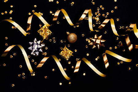 Decorative present boxes, balls, golden streamer, decorative bows, stars and sequins on black background. Party and festive concept. Top view. Flat lay.