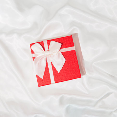 Red present box with bow on white silk sheet. Christmas or birthday gift. Festive concept. Top view. Flat layの写真素材