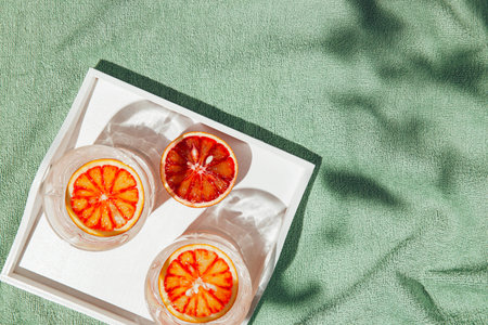 Summer scene with half of bloody orange glasses of water with white wooden tray on pastel green...