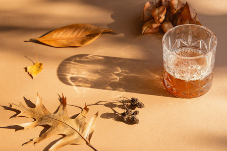 Glass with homemade drink on light brown sunlit background with dried leaves and shadows. Autumn and fall conceptの写真素材