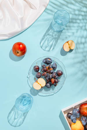 Creative composition made of plate with fruits, glasses, apples on pastel blue sunlit background with white silk cloth and palm tree leaves shadows. Summer and refreshment concept. Harvest themeの写真素材