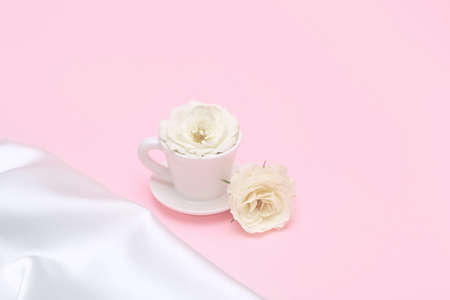 Creative composition made of white cup of coffee or tea with rose flower bud and silk cloth on pastel pink background. Morning beverage concept. Minimal styleの写真素材
